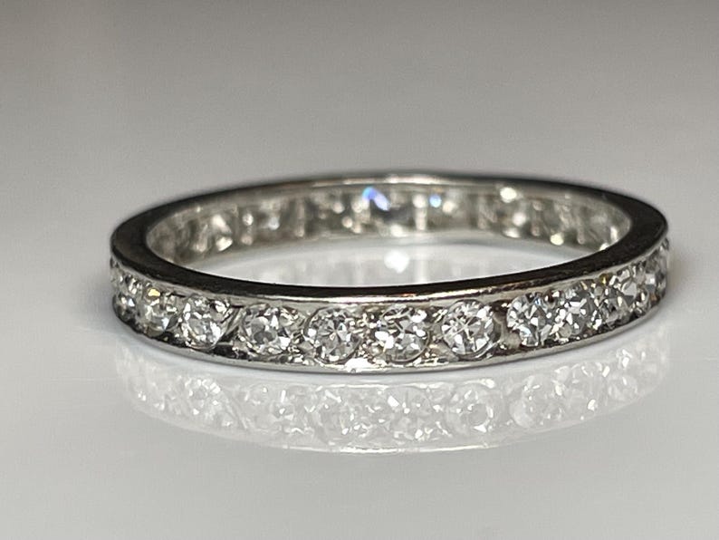 May include: A silver eternity ring featuring a continuous row of round-cut diamonds. The diamonds are set closely together, creating a sparkling band. The ring is displayed on a reflective surface.