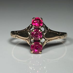 May include: A gold-toned ring featuring three vibrant, round, red gemstones. The ring has an ornate, openwork design with delicate etched details on the band. The gemstones are set in a vertical row, creating a classic and elegant look.