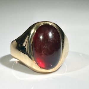 Könnte beinhalten: Ein goldener Ring mit einem großen, ovalen, dunkelroten Edelstein. Der Ring hat ein klassisches Siegelring-Design mit einem polierten Goldband. Der Edelstein ist glatt und bündig mit der Oberfläche des Rings.