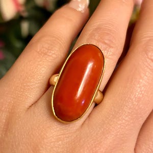 May include: A gold ring featuring a large, oval-shaped, polished red stone. The stone is set in a gold band, creating a classic and elegant design. The ring is shown on a hand, highlighting its size and color.