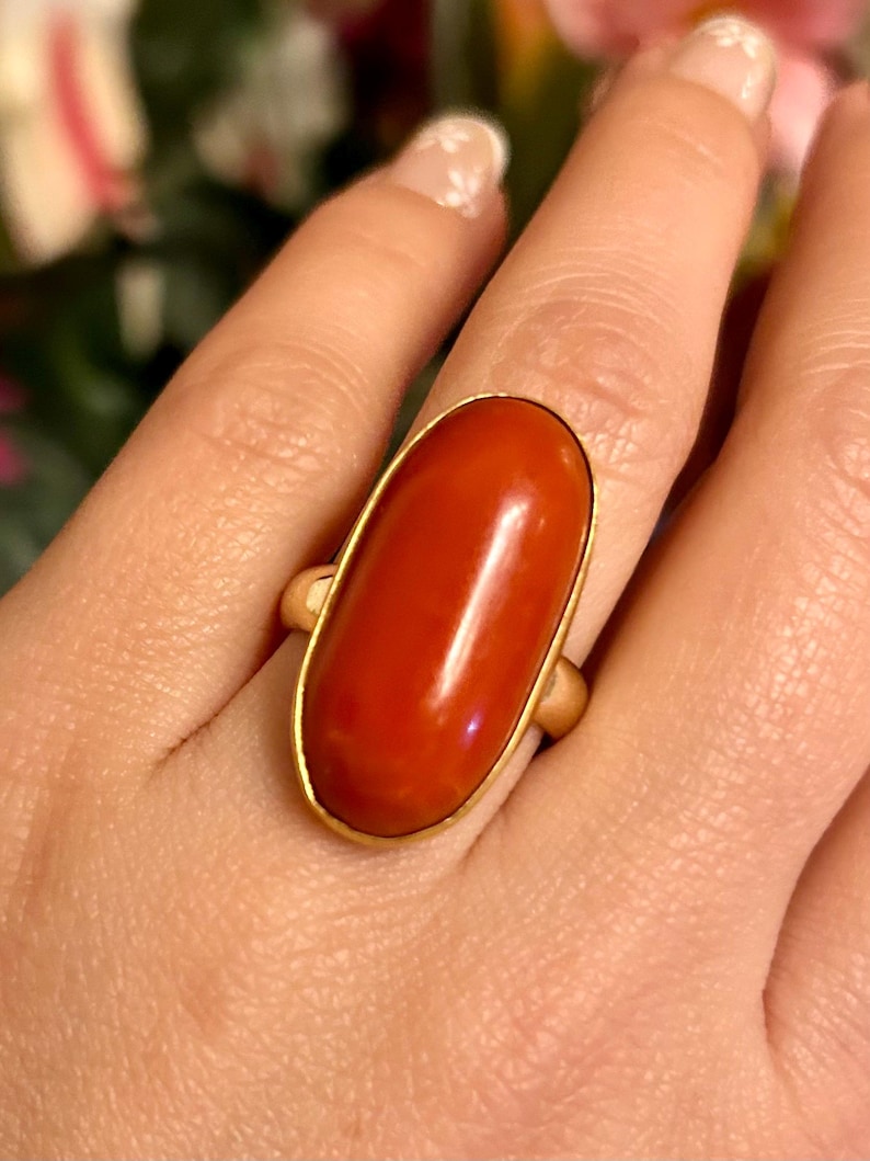 May include: A gold ring featuring a large, oval-shaped, polished red stone. The stone is set in a gold band, creating a classic and elegant design. The ring is shown on a hand, highlighting its size and color.