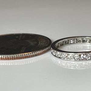 May include: A silver eternity ring with numerous small, clear gemstones set around the band. The ring is next to a U.S. quarter coin for size comparison. The ring is on a white surface.