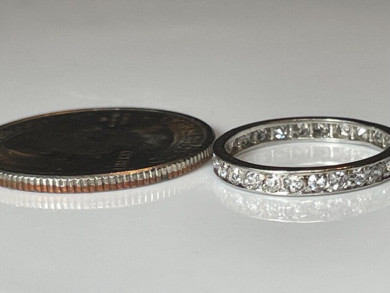 May include: A silver eternity ring with numerous small, clear gemstones set around the band. The ring is next to a U.S. quarter coin for size comparison. The ring is on a white surface.