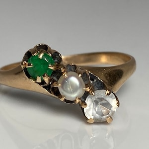 Puede incluir: Un anillo de oro con tres piedras: una esmeralda verde, una perla blanca y una piedra transparente. Las piedras están engastadas en garras.