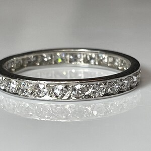 May include: A silver ring featuring a continuous row of small, clear gemstones. The ring's band is set with multiple round-cut stones, creating a sparkling effect. The ring is shown on a reflective surface.