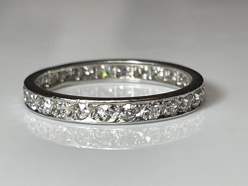 May include: A silver ring featuring a continuous row of small, clear gemstones. The ring's band is set with multiple round-cut stones, creating a sparkling effect. The ring is shown on a reflective surface.