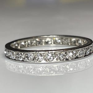 May include: A silver band ring featuring a continuous line of clear gemstones. The ring is displayed on a reflective surface, accentuating the sparkle of the stones. The design is simple and elegant, suitable for various occasions.