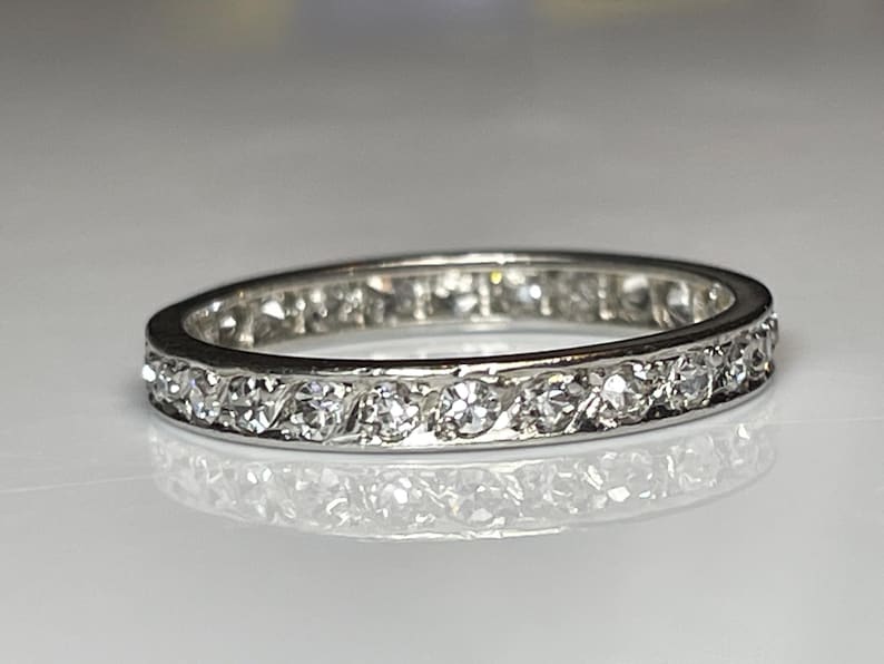 May include: A silver band ring featuring a continuous line of clear gemstones. The ring is displayed on a reflective surface, accentuating the sparkle of the stones. The design is simple and elegant, suitable for various occasions.