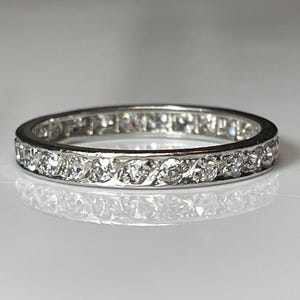 May include: A silver ring featuring a continuous band of small, clear gemstones. The ring is circular and rests on a reflective surface, with the gemstones set closely around the band. The ring's design is simple and elegant, suitable for everyday wear.