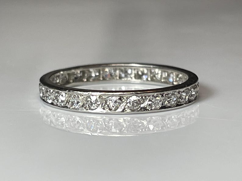 May include: A silver ring featuring a continuous band of small, clear gemstones. The ring is circular and rests on a reflective surface, with the gemstones set closely around the band. The ring's design is simple and elegant, suitable for everyday wear.