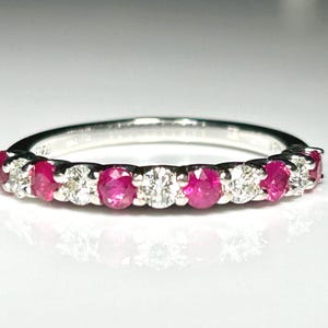 May include: A silver ring featuring a row of alternating round-cut rubies and diamonds. The gemstones are set closely together, creating a continuous band of color and sparkle. The ring is displayed on a white surface.