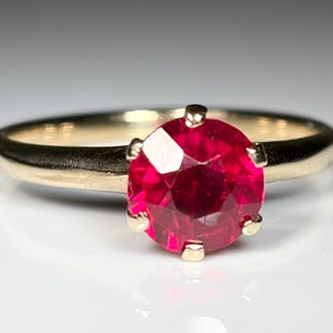 Puede incluir: Un anillo de oro con una piedra preciosa roja vibrante, redonda y facetada, engastada en una montura de seis garras. La banda es lisa y pulida, mostrando un diseño clásico con un acabado brillante que refleja la luz sobre el metal.