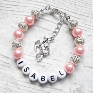 May include: A pink beaded bracelet with the name "ISABEL" spelled out in white letter beads. The bracelet features silver-colored, textured beads and a unicorn charm. The bracelet is designed for a child.