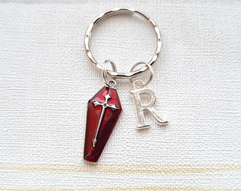 May include: A silver key chain with a red coffin charm and a silver letter R charm. The coffin charm has a silver cross on it.