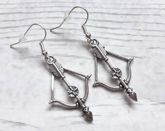 May include: A pair of silver-toned dangle earrings featuring a bow and arrow design. Each earring has a metal bow frame with a central arrow, complete with fletching details and a pointed tip, suspended from a standard silver-toned hook wire.