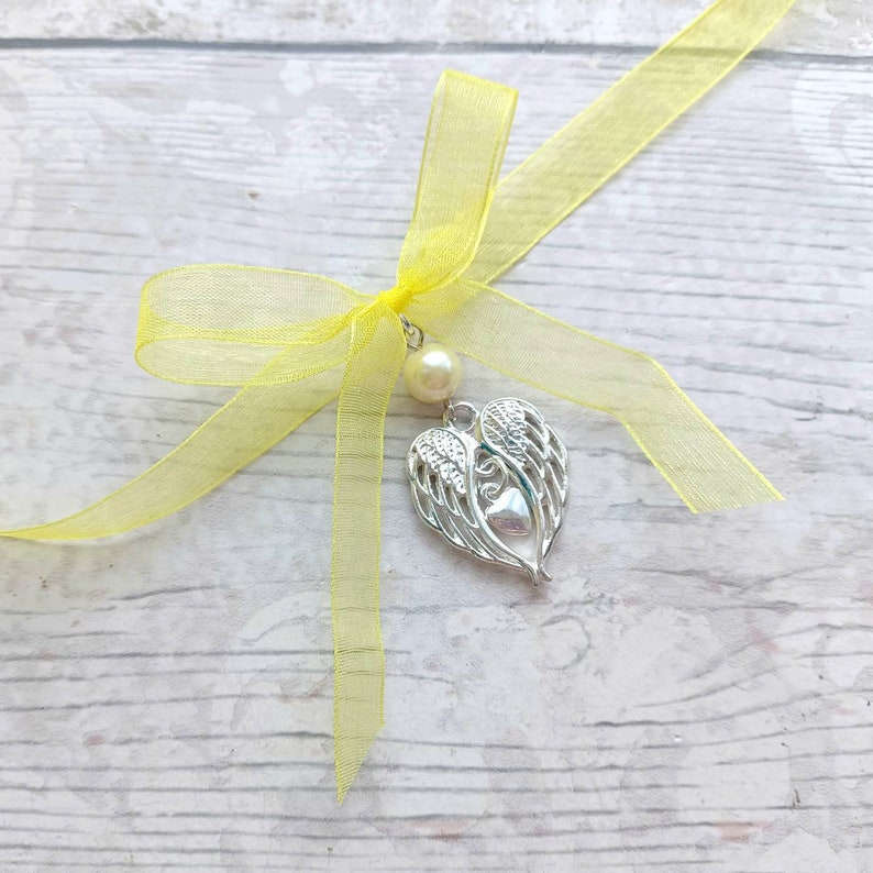 May include: A yellow ribbon tied into a bow with a silver angel wing charm and a pearl bead hanging from the center.