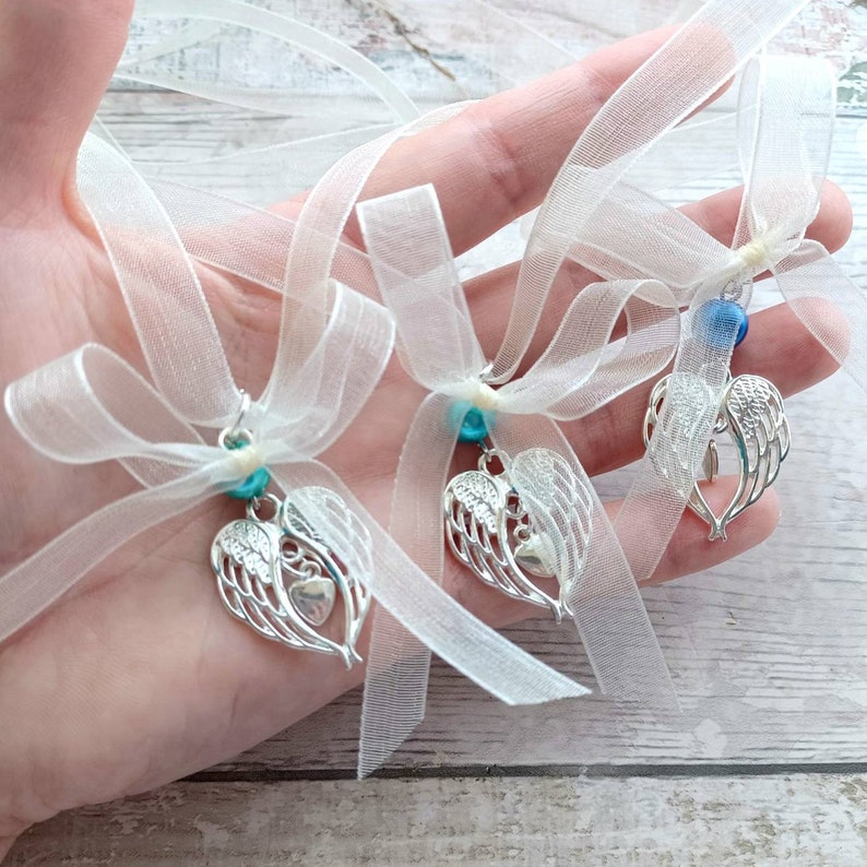 May include: Three white tulle ribbon favors with silver angel wing charms and blue beads. The charms are shaped like a heart with wings.