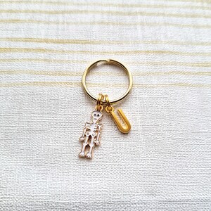 以下が含まれることがあります： 金色のスケルトンキーホルダーに金色のUの文字のチャームが付いている。