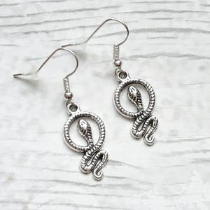 Puede incluir: Pendientes de serpiente plateados con un diseño de serpiente enroscada. Los pendientes son de estilo colgante con un gancho plateado.