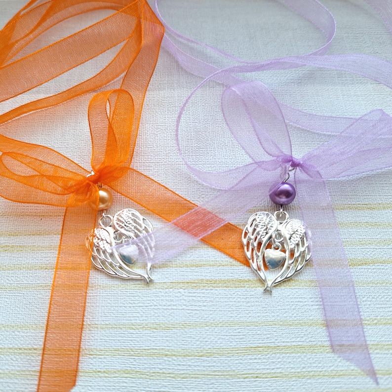 May include: Two angel wing heart charms with a pearl bead on a ribbon. One ribbon is orange and the other is lavender.