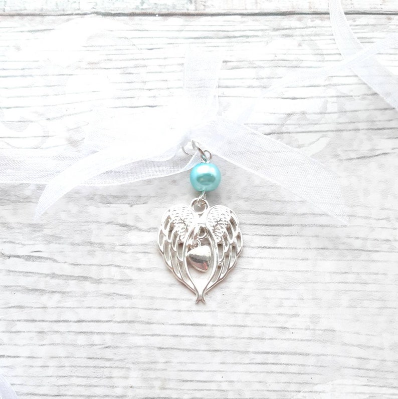 May include: A silver charm with angel wings and a heart pendant, attached to a white ribbon with a light blue pearl bead.