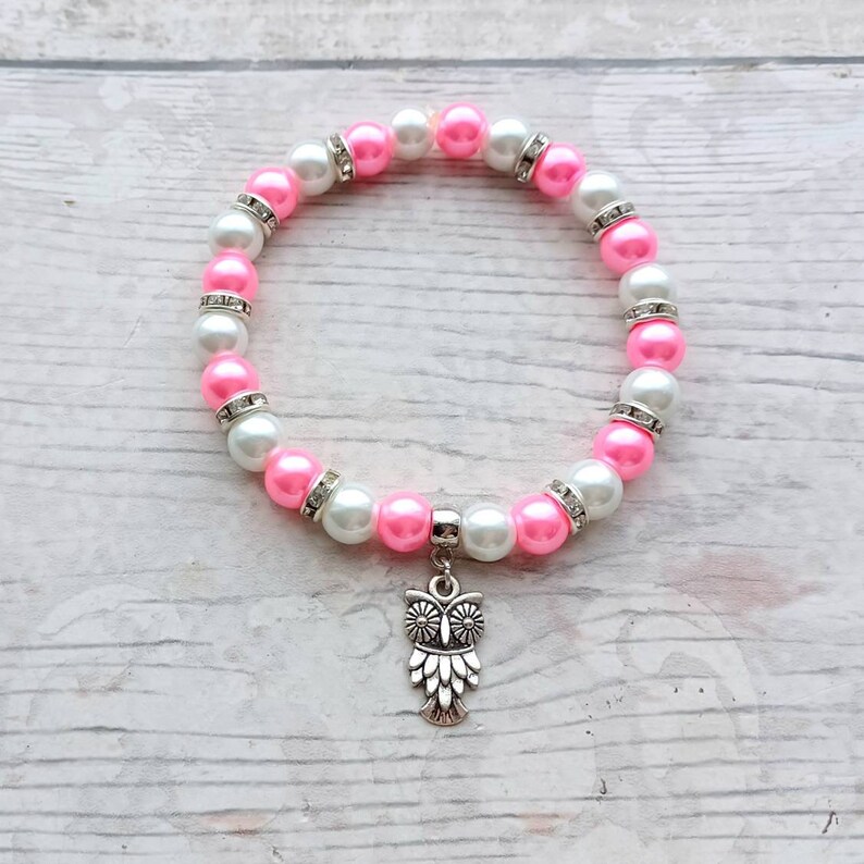 May include: A pink and white beaded bracelet with a silver owl charm. The beads are alternating pink and white with rhinestones.