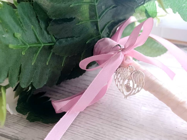 May include: A white and green bouquet with a pink ribbon and a silver charm with angel wings and a heart.