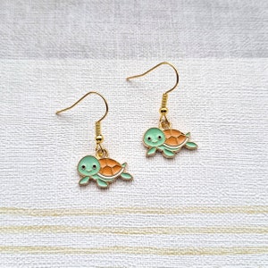 May include: A pair of gold dangle earrings featuring a green and orange turtle charm. The turtle has a smiling face and is swimming to the right.