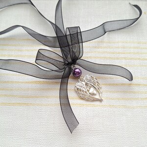 May include: A black ribbon tied into a bow with a silver heart charm with angel wings and a purple pearl hanging from it.