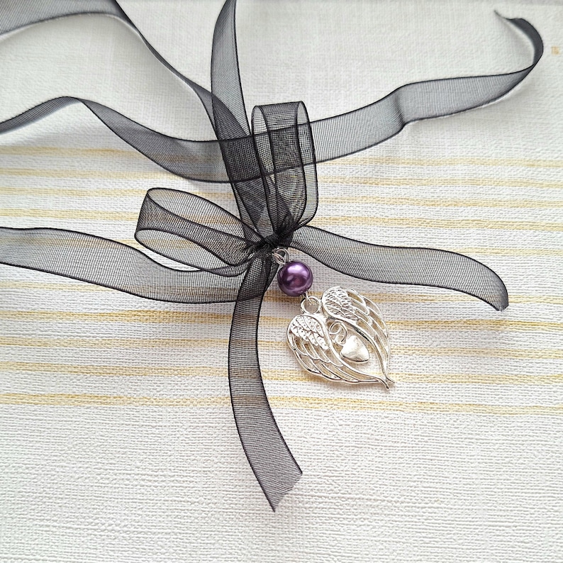 May include: A black ribbon tied into a bow with a silver heart charm with angel wings and a purple pearl hanging from it.