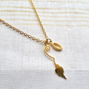 Puede incluir: Un collar dorado con una cadena delicada. El colgante presenta un diseño de hoja estilizada y un pequeño dije ovalado. El collar se muestra sobre una superficie texturizada blanquecina.