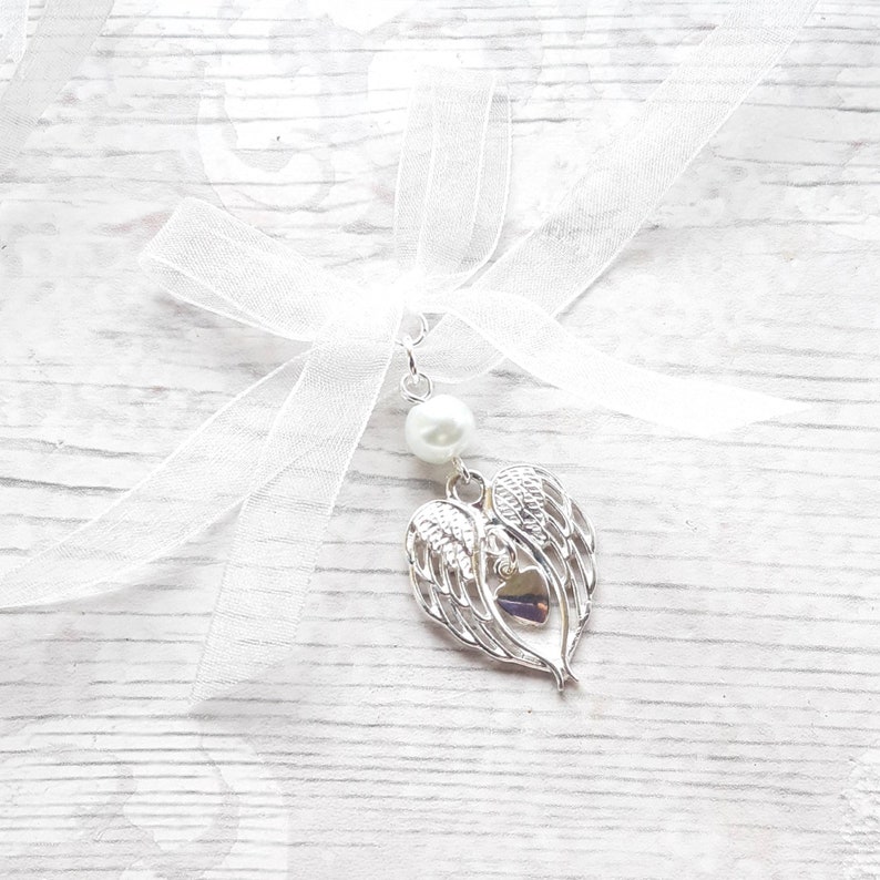 May include: A white sheer ribbon with a silver angel wing charm and a pearl bead. The charm features a heart with wings.
