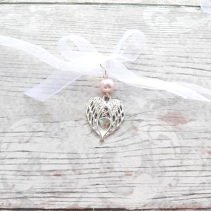 May include: A silver charm with angel wings and a heart in the center, attached to a white ribbon with a pink bead.