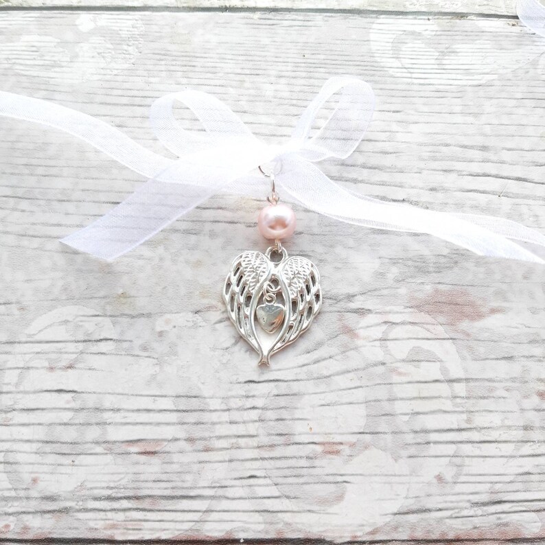 May include: A silver charm with angel wings and a heart in the center, attached to a white ribbon with a pink bead.