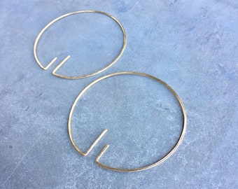 May include: Two gold-colored hoop earrings with a unique design. Each earring features a circular shape with a small, angular extension on one side. The earrings are displayed on a gray surface.