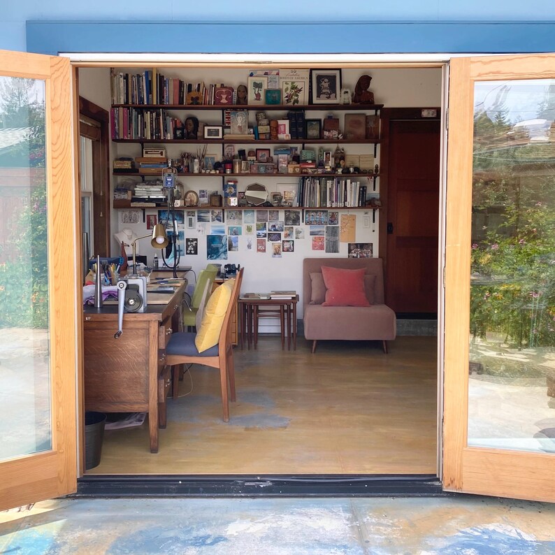 May include: An interior shot of a home office or studio, viewed through open double doors. The room features a wooden desk, bookshelves filled with books and decor, a small seating area, and a wall covered in photos and artwork. The space is well-lit and inviting.
