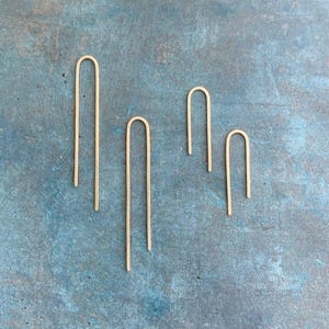 May include: Four gold-colored hair pins of varying lengths are arranged on a textured blue surface. The pins have a simple U-shape design. The pins are made of a shiny metal and are evenly spaced apart.
