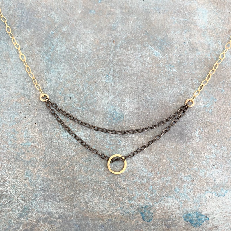 May include: A delicate necklace featuring a gold chain and two layers of dark gray chains. A small gold circle pendant hangs from the bottom chain. The necklace is displayed against a textured, gray-blue surface.