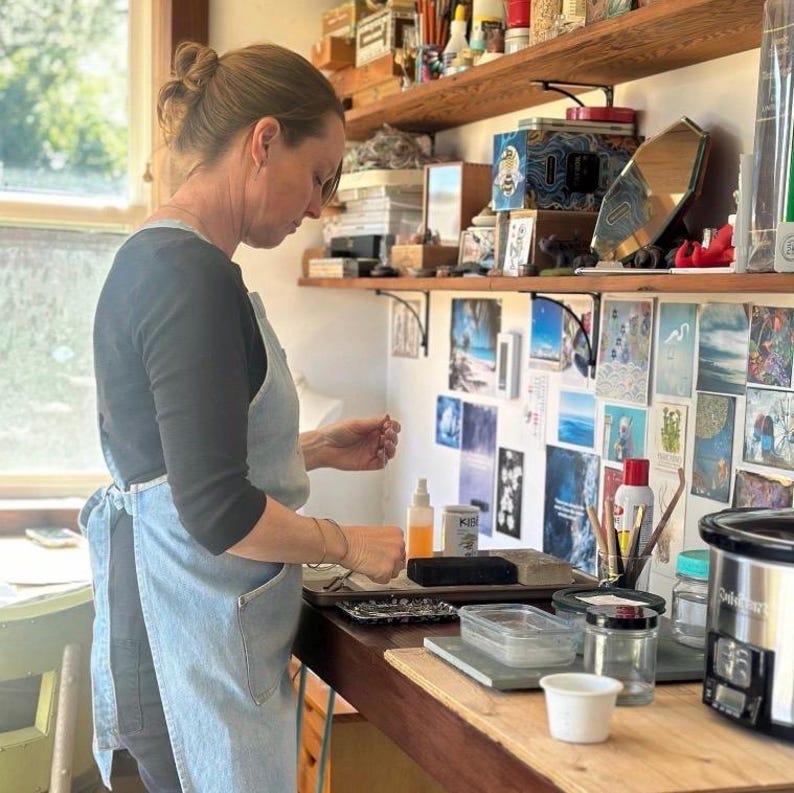 May include: A person wearing a light blue apron and black long-sleeve shirt working in a well-lit workshop. The workspace includes shelves with various items, a window, and a wooden countertop with tools and supplies.