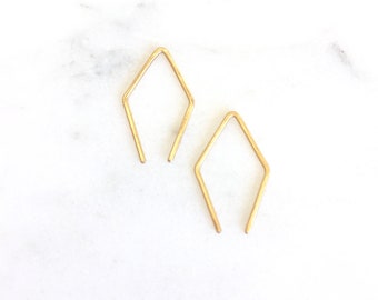 May include: Two delicate, gold-colored earrings. Each earring is a geometric open diamond shape with a straight post at the bottom. The earrings are displayed on a white marble surface.
