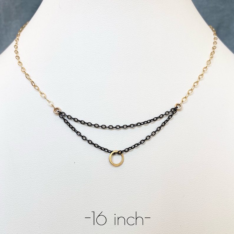 May include: A delicate necklace with a two-tiered chain design. The top chain is gold-colored, transitioning to a black chain below. A small gold-colored circle pendant hangs from the bottom chain. The necklace is approximately 16 inches long.