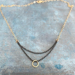 May include: A layered necklace with a gold chain and a dark gray chain. The necklace features a small gold circle pendant. The background is a textured blue-gray surface. The necklace is a piece of jewelry.