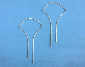 Może przedstawiać: Para złotych kolczyków w kształcie wachlarza na niebieskim tle. Kolczyki mają delikatny, minimalistyczny design, z zakrzywioną górą i dwiema prostymi liniami biegnącymi w dół.