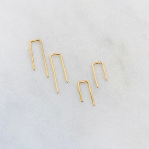 May include: Four gold-colored, open-ended, U-shaped hoops of varying sizes. The hoops are arranged in a staggered pattern on a white surface.