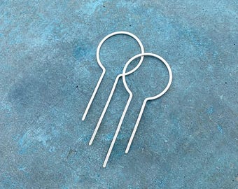 Könnte beinhalten: Zwei Paar Ohrringe aus weißem Metall. Jeder Ohrring hat einen kreisförmigen oberen Teil und einen geraden, länglichen Stab. Die Ohrringe sind vor einem strukturierten, blau-grünen Hintergrund platziert.