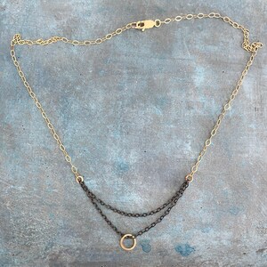 May include: A gold-toned necklace with a delicate chain and a layered design. The upper chain is gold, while the lower layers are a darker, contrasting color, with a small gold circle pendant. The necklace is displayed on a blue-grey surface.