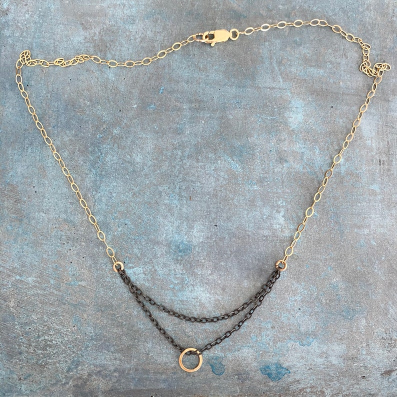 May include: A gold-toned necklace with a delicate chain and a layered design. The upper chain is gold, while the lower layers are a darker, contrasting color, with a small gold circle pendant. The necklace is displayed on a blue-grey surface.