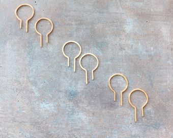 May include: Six gold-colored, open-circle earrings with a small, straight post extending from the bottom. The earrings are arranged in pairs on a mottled, gray-blue surface. The earrings have a simple, minimalist design.