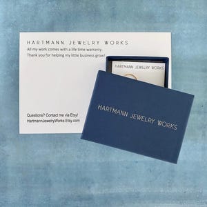 May include: A navy blue jewelry box with the text "HARTMANN JEWELRY WORKS" in silver lettering. The box is open, revealing a small gold ring. A white card with the same text and a lifetime warranty statement is also visible.