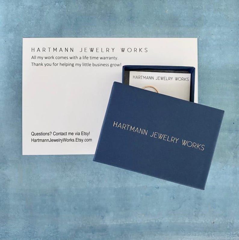 May include: A navy blue jewelry box with the text "HARTMANN JEWELRY WORKS" in silver lettering. The box is open, revealing a small gold ring. A white card with the same text and a lifetime warranty statement is also visible.